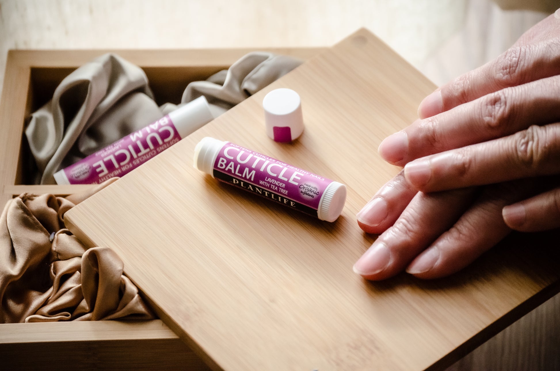 Plantlife Lavender Tea Tree Cuticle Balm being applied to nails, shown on a wooden tray with soft fabric and packaging in the background.