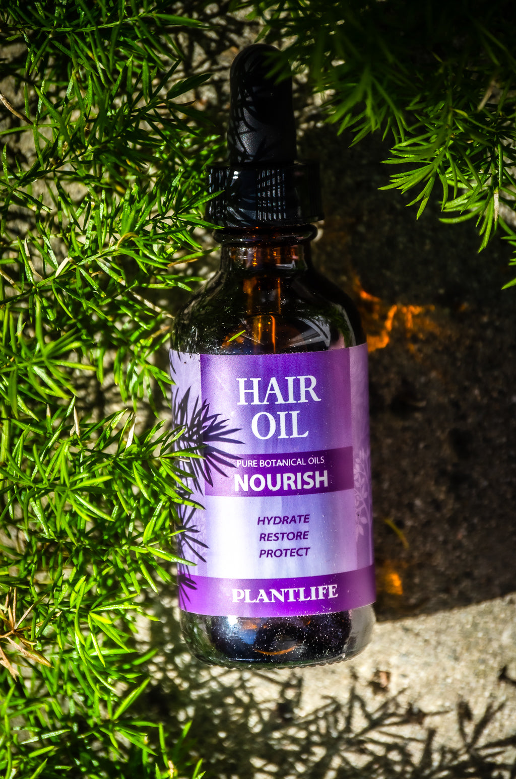 Plantlife Nourish Hair Oil bottle lying on a stone surface surrounded by green foliage in natural outdoor lighting.