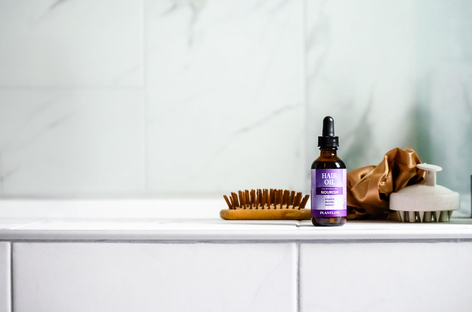 Plantlife Nourish Hair Oil on a bathroom ledge with a wooden brush, scalp massager, and silk scrunchie in frame.