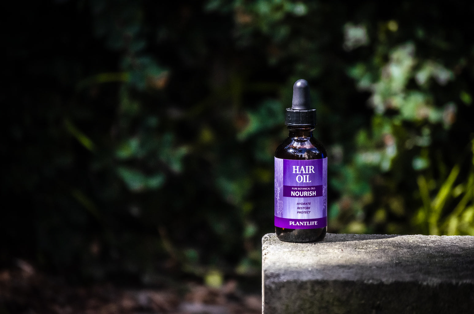 Plantlife Nourish Hair Oil bottle placed outdoors on a stone surface with green foliage background.