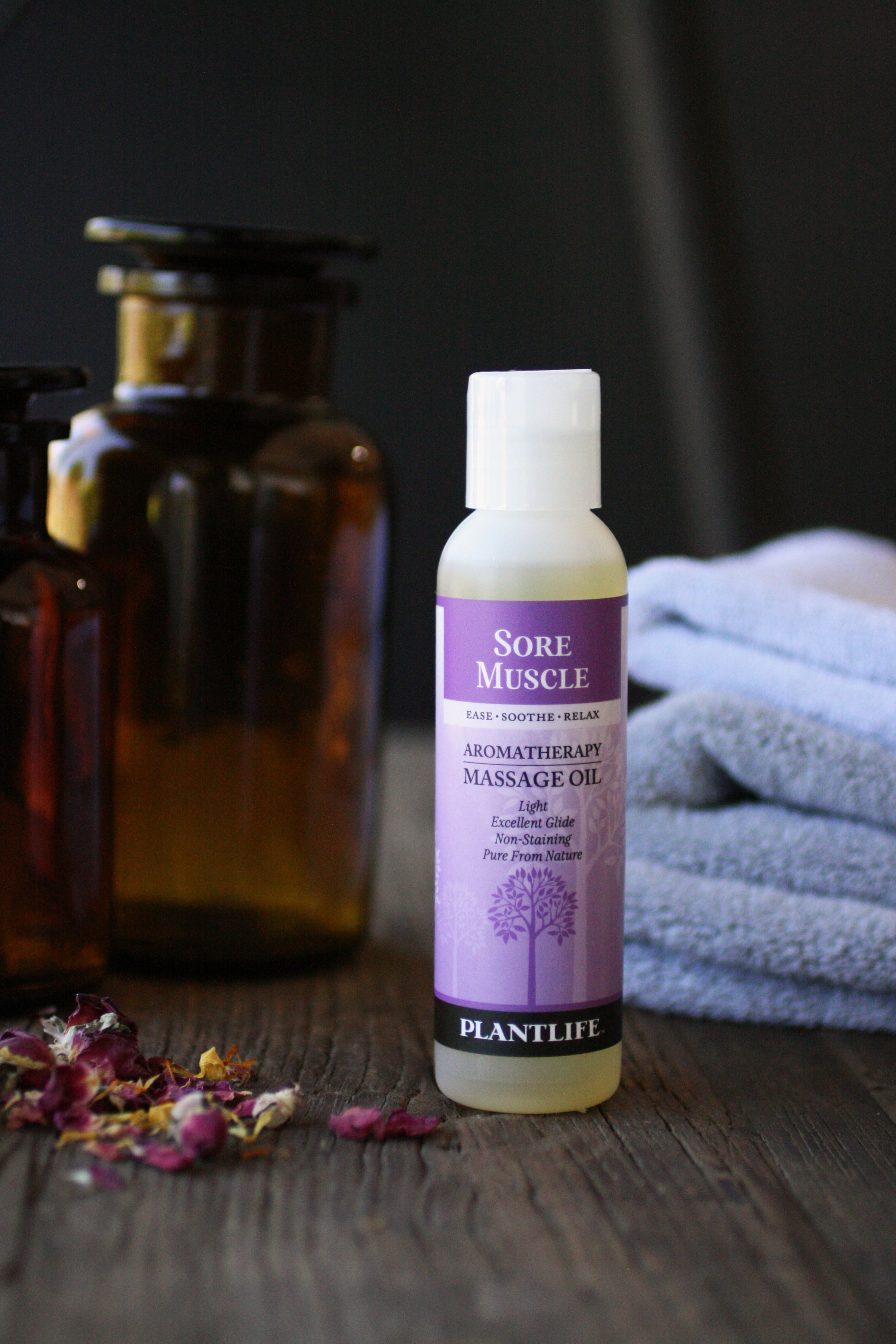 Bottle of Plantlife Sore Muscle massage oil on a wooden surface with towels and bottles in the background.
