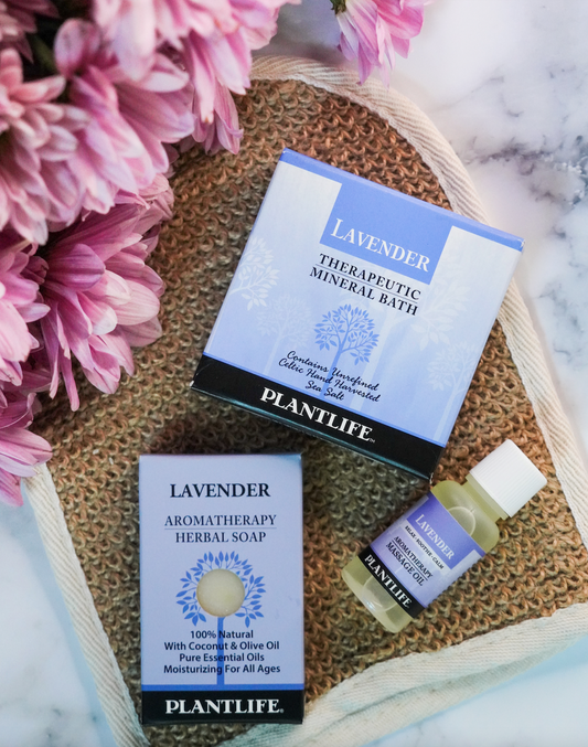 Plantlife Lavender gift set with therapeutic mineral bath, aromatherapy herbal soap, and massage oil, styled with purple flowers on a natural fiber mat