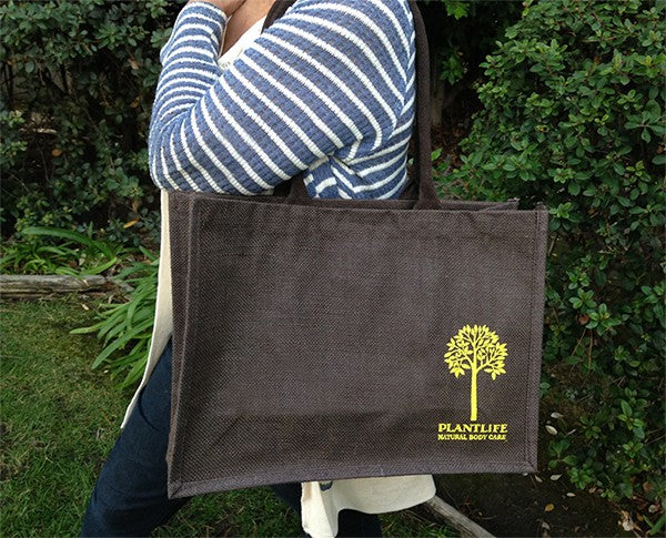 Person holding a dark brown Plantlife Natural Body Care Jute Tote Bag with yellow tree logo, featuring long handles and durable woven material, shown outdoors.