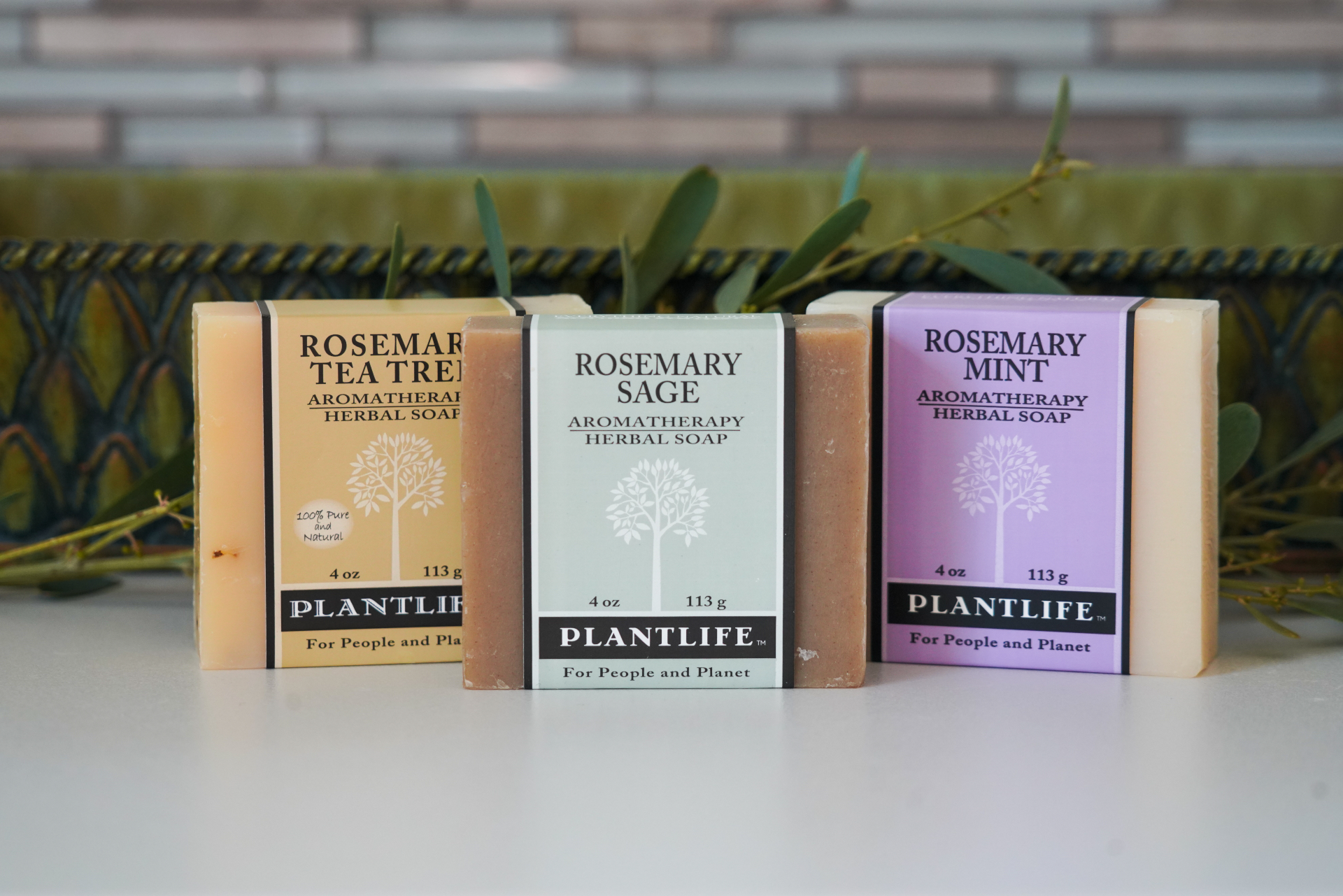 Plantlife Rosemary Tea Tree, Rosemary Sage, and Rosemary Mint aromatherapy herbal soaps displayed in a row with greenery and decorative backdrop