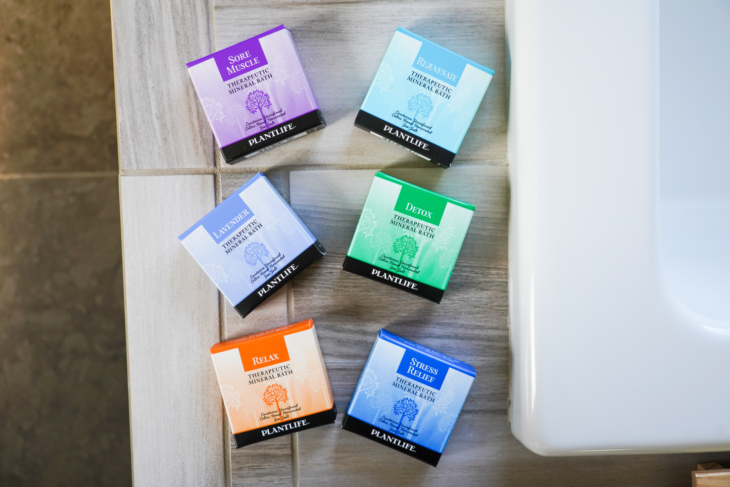 Flat lay of six Plantlife Therapeutic Mineral Bath boxes—Sore Muscle, Rejuvenate, Lavender, Detox, Relax, and Stress Relief—arranged diagonally on gray tile