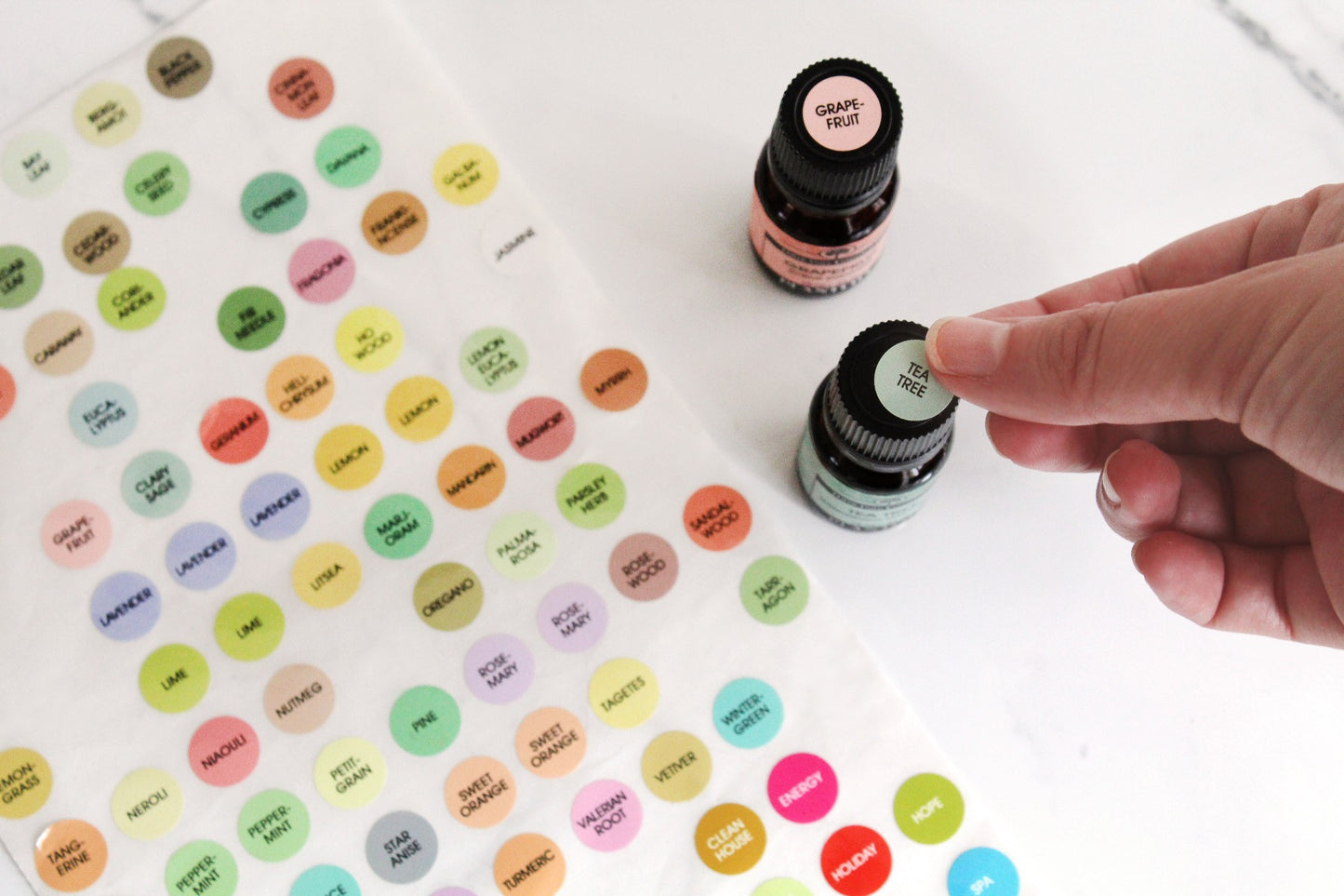 A person is applying a color-coded "TEA TREE" essential oil label to the top of an amber essential oil bottle. A "GRAPEFRUIT" labeled bottle is also shown nearby. A full sheet of Plantlife’s round, colorful oil identification stickers with various oil names is spread on the table.