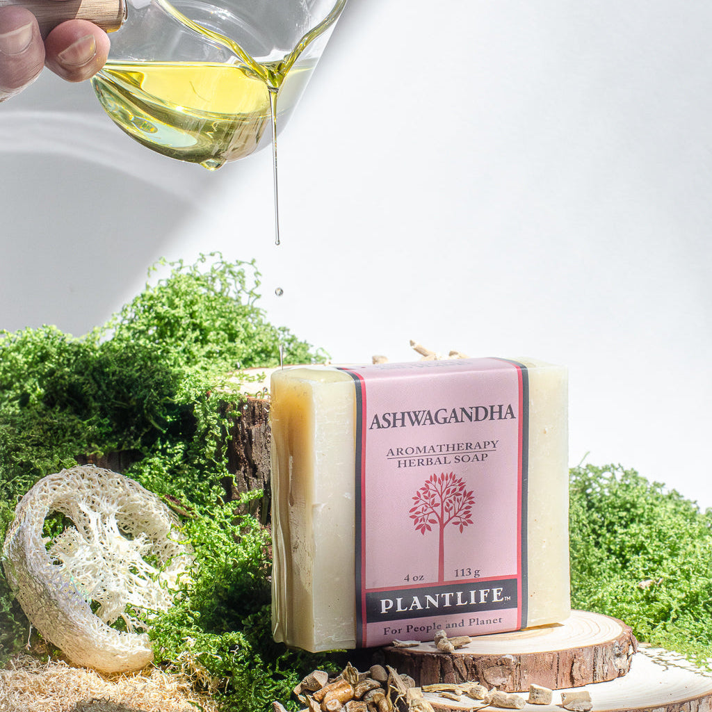 Plantlife Ashwagandha Aromatherapy Herbal Soap bar in a pink wrapper, styled with a lathered soap sponge, dried herbs, moss, and a hand pouring oil from a glass pitcher.