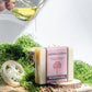 Plantlife Ashwagandha Aromatherapy Herbal Soap bar in a pink wrapper, styled with a lathered soap sponge, dried herbs, moss, and a hand pouring oil from a glass pitcher.