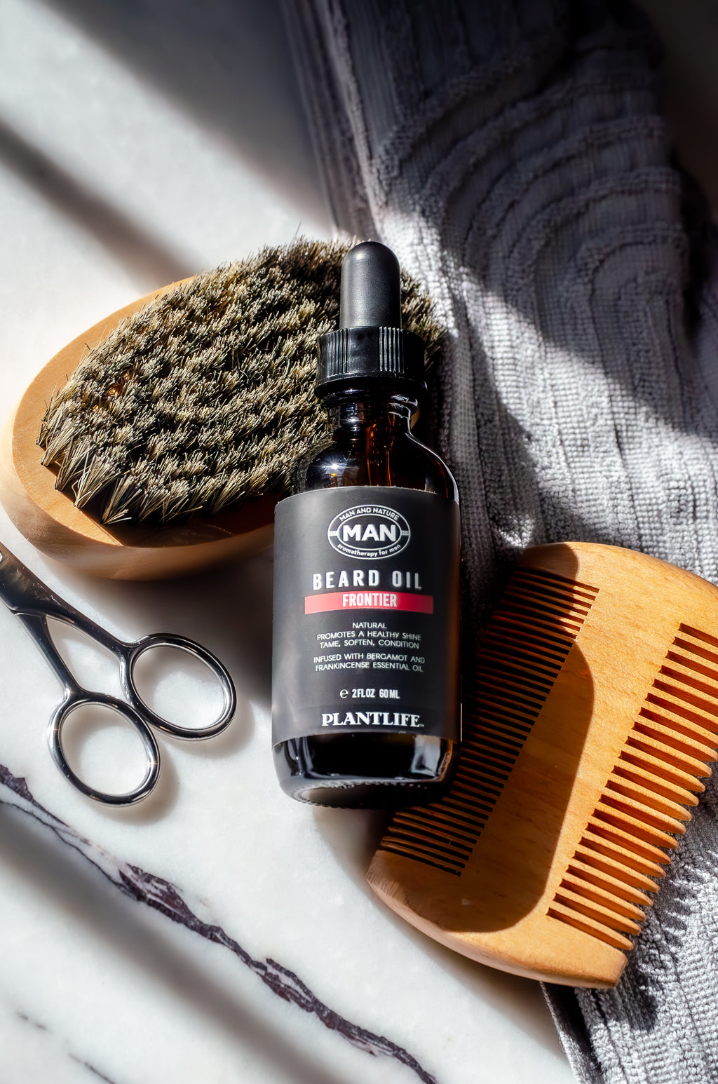 Plantlife Frontier Beard Oil 2 oz with wooden beard brush, comb, and grooming scissors arranged on a marble surface