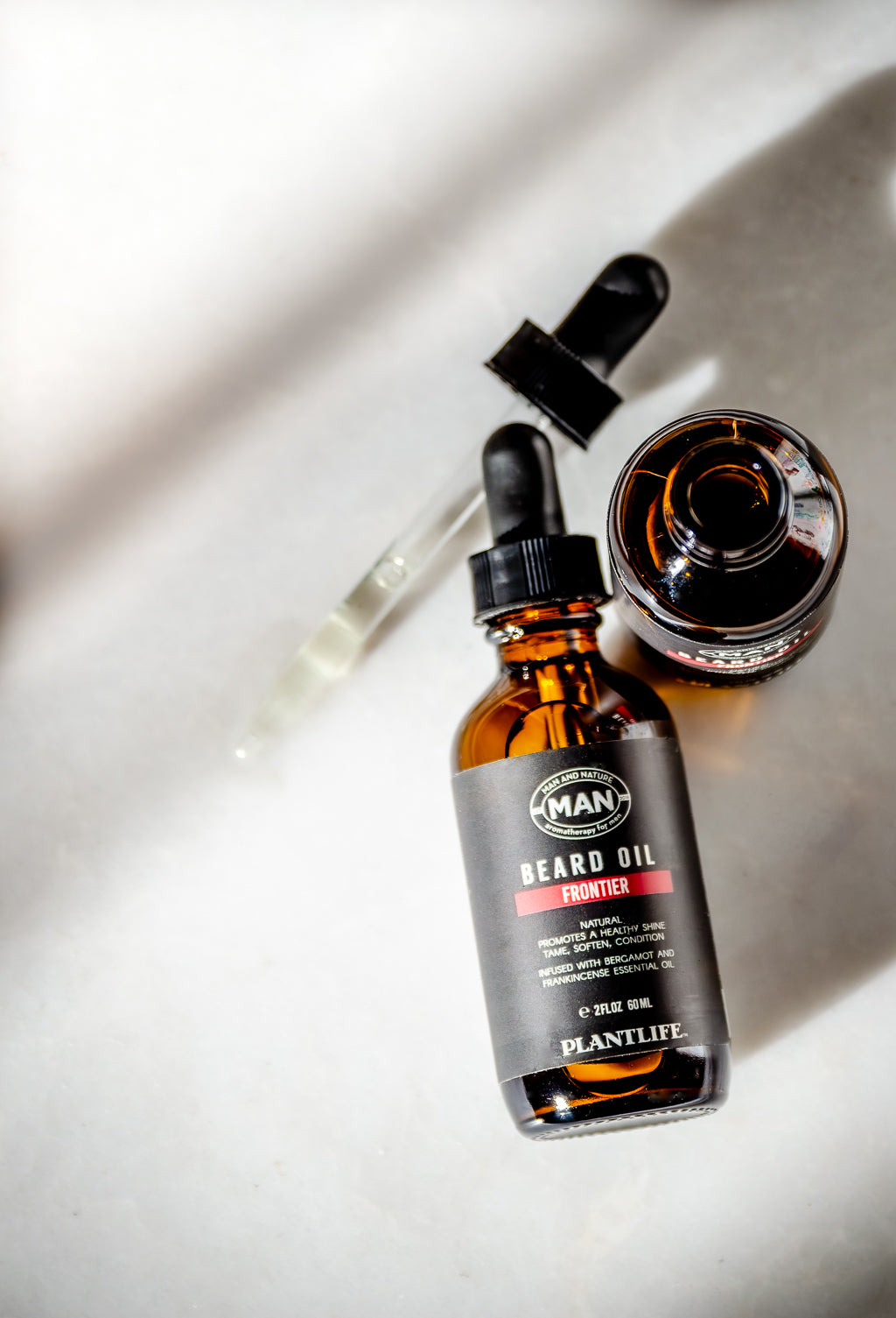 Plantlife Man Beard Oil Frontier in an amber glass dropper bottle with a black cap, shown next to an open bottle and a glass dropper on a light gray surface with dramatic shadows.