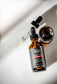 Plantlife Man Beard Oil Frontier in an amber glass dropper bottle with a black cap, shown next to an open bottle and a glass dropper on a light gray surface with dramatic shadows.