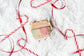 A packaged bar soap set placed on a white textured surface with red-and-white candy cane ribbons around it. The package reads Suds of the Season and 'Holiday Bar Soap,' with three visible soap bars in pastel green, pink, and peach tones.