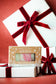 Plantlife Christmas bar soap in a kraft paper box with a clear window, showing pink and green bars inside, styled on top of white gift boxes with red velvet ribbons against a red background.