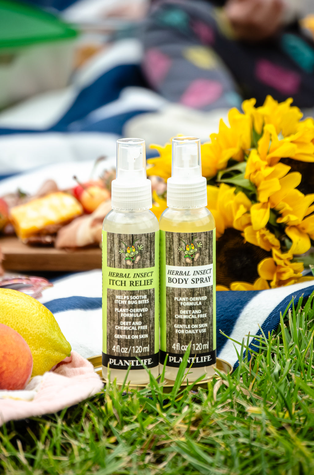 Plantlife Herbal Insect Itch Relief and Body Spray 4 fl oz bottles displayed outdoors at a picnic with flowers, fruit, and grass
