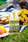 Plantlife Herbal Insect Itch Relief and Body Spray 4 fl oz bottles displayed outdoors at a picnic with flowers, fruit, and grass