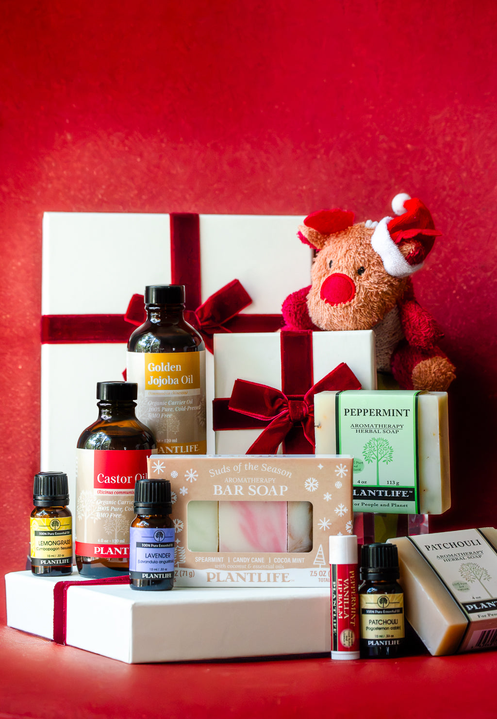 A Christmas-themed gift set featuring various bath and body products including Plantlife bar soap, Castor Oil, Golden Roghan Oil, and essential oils, arranged with white gift boxes tied with red ribbons and a red plush reindeer toy against a textured red background.