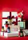 A Christmas-themed gift set featuring various bath and body products including Plantlife bar soap, Castor Oil, Golden Roghan Oil, and essential oils, arranged with white gift boxes tied with red ribbons and a red plush reindeer toy against a textured red background.
