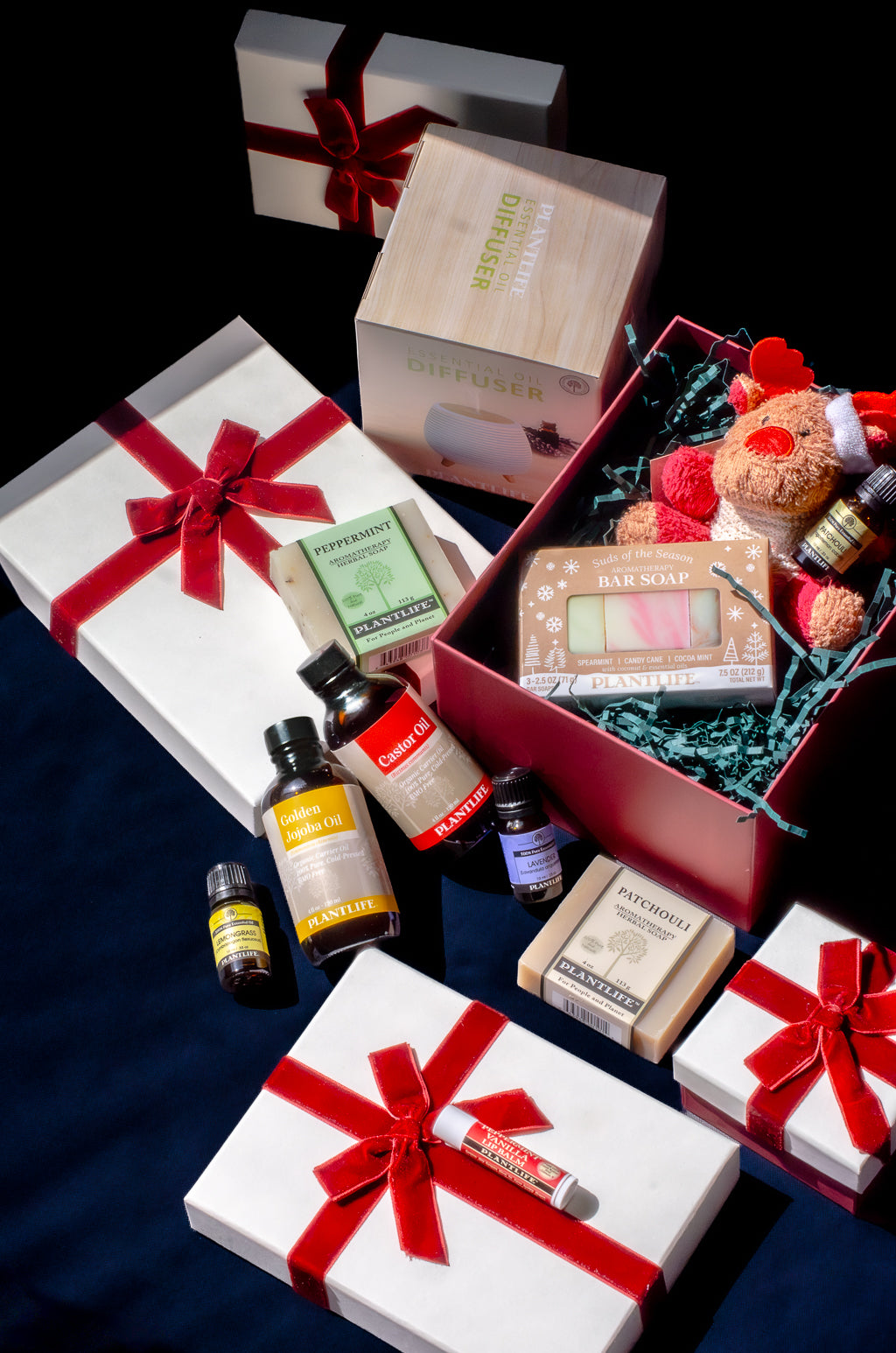 A curated gift set arranged on a dark background, featuring multiple white gift boxes tied with red ribbons, an open red gift box filled with teal crinkle paper and a small teddy bear, bottles of essential oils, a boxed soap, and a lip balm tube.