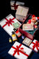 A curated gift set arranged on a dark background, featuring multiple white gift boxes tied with red ribbons, an open red gift box filled with teal crinkle paper and a small teddy bear, bottles of essential oils, a boxed soap, and a lip balm tube.
