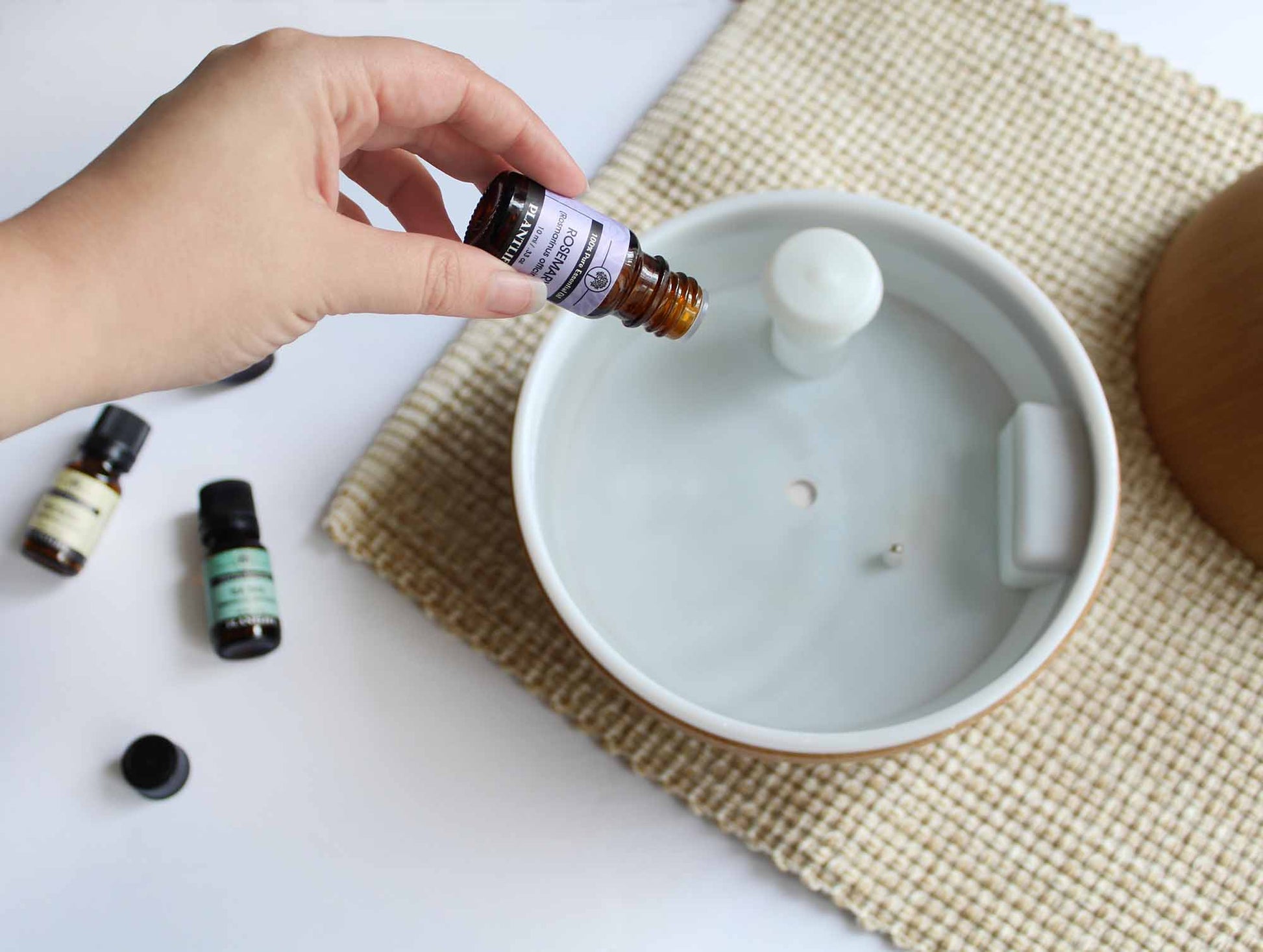 Hand pouring essential oil into a diffuser on a white surface with a textured mat.