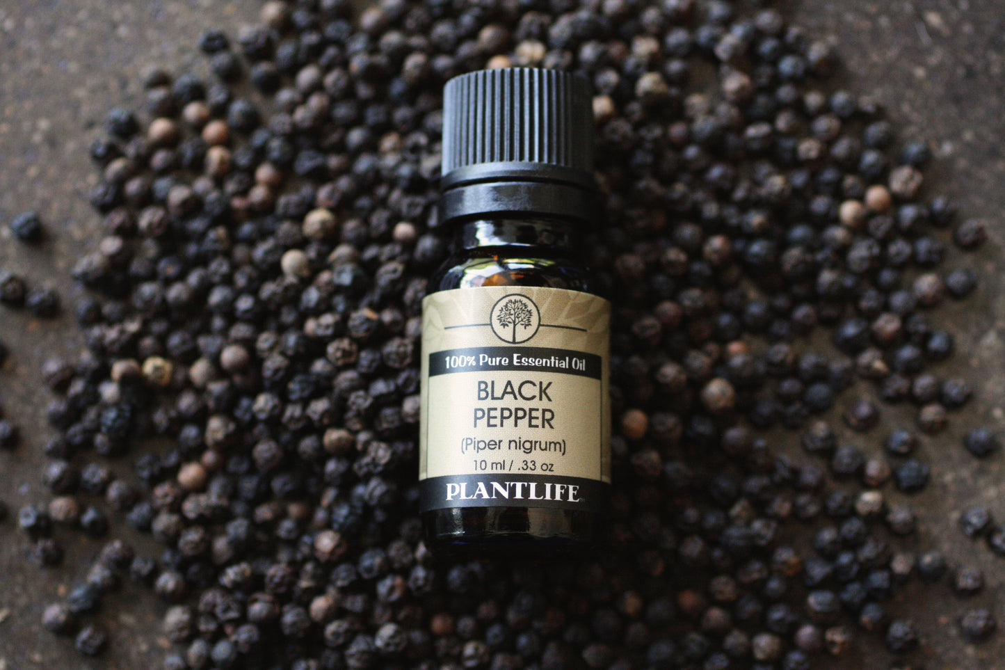Bottle of Plantlife Black Pepper essential oil surrounded by black peppercorns on a dark surface