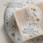 Close-up of a creamy beige Plantlife soap bar resting in foamy water. Text on the image highlights “Made with patchouli essential oil to help soothe and refresh the skin” and “Rich lather, moisturizing and gentle on skin.”