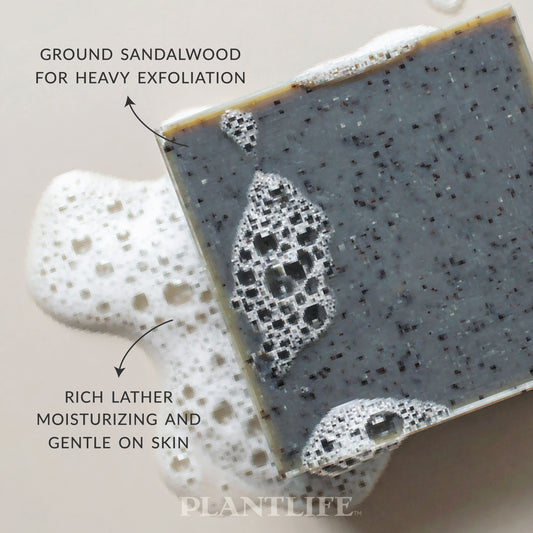 Close-up of a dark gray Plantlife soap bar with visible exfoliating particles resting in foamy water. Text on the image highlights “Ground sandalwood for heavy exfoliation” and “Rich lather, moisturizing and gentle on skin.”