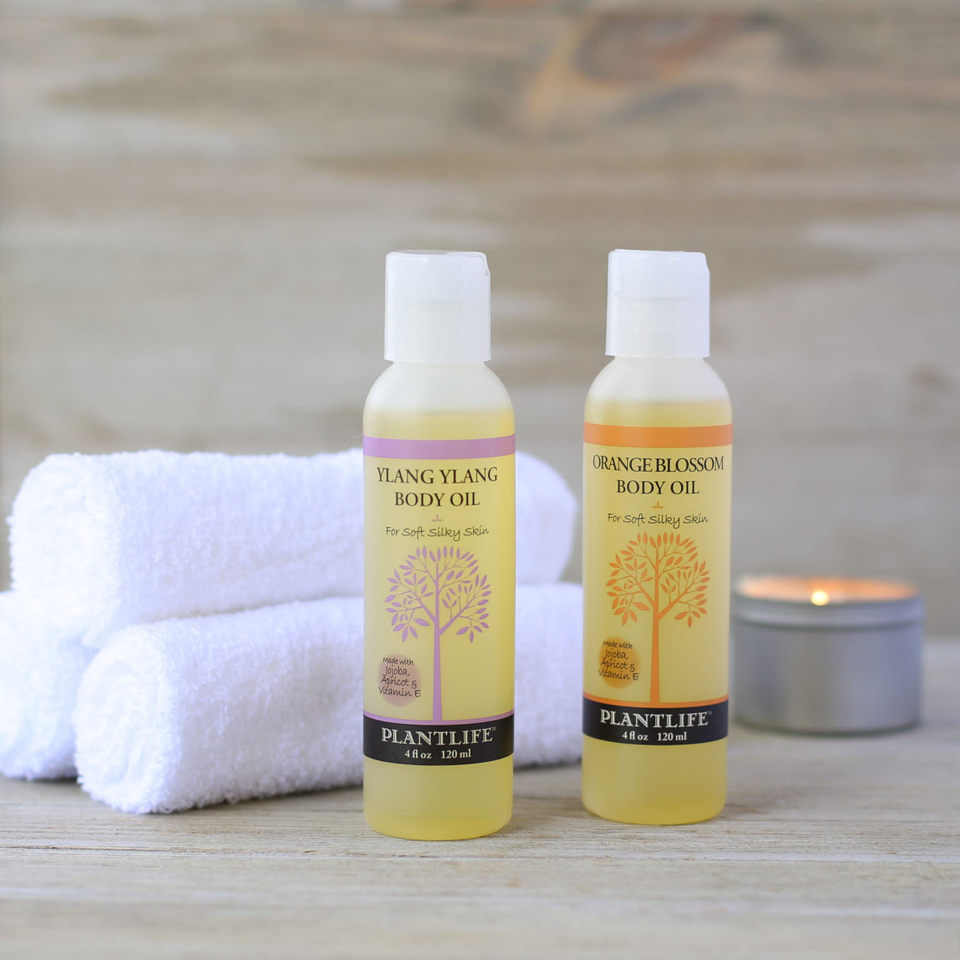 Two bottles of Plantlife body oil on a wooden surface with towels and a candle in the background.