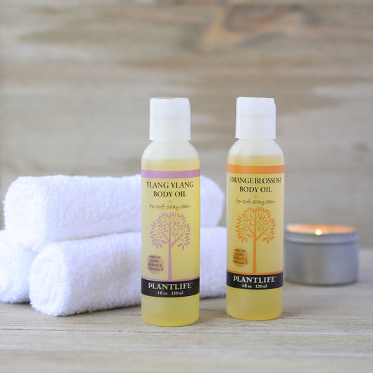 Two bottles of Plantlife body oil on a wooden surface with towels and a candle in the background.